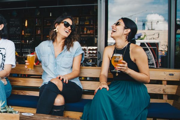 Two friends sitting side by side on a bench, holding amber glasses of beer and laughing.
